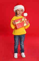 Cute little girl in Santa hat with Christmas gift on red background