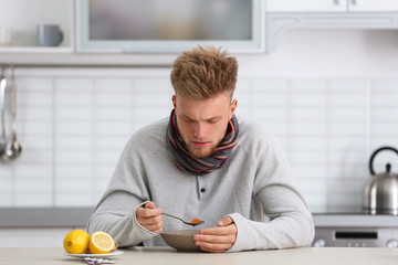 Sick young man eating soup to cure flu at table in kitchen