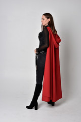 full length portrait of a pretty brunette woman wearing black leather fantasy costume with long red superhero cape. standing pose on a studio background.