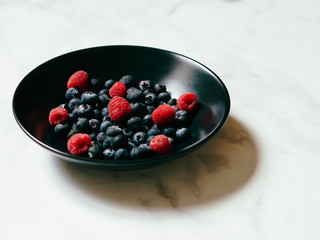 Blueberries and Raspberries in a black dish on a white marble background
