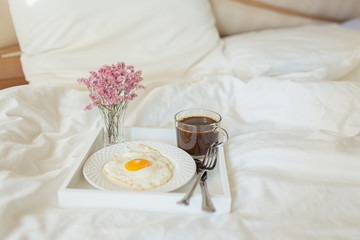 White tray with breakfast on a bed in a hotel room. Fried egg, cup of coffee and flowers in white sheets in light bedroom.