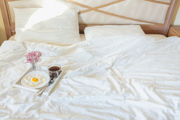 Obraz na płótnie Canvas White tray with breakfast on a bed in a hotel room. Fried egg, cup of coffee and flowers in white sheets in light bedroom. Copyspace.