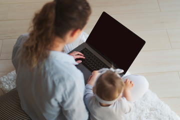 Back view.Mother and daughter sitting on floor and looking at screen of laptop. Woman freelance worker trying to do her job.