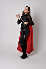 full length portrait of a pretty brunette woman wearing black leather fantasy costume with long red superhero cape. standing pose on a studio background.
