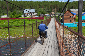 A motorcyclist rides an off-road motorcycle on a suspended wooden bridge over a mountain river.