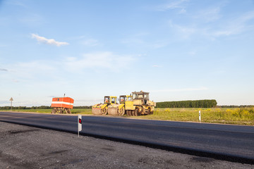 Three large yellow asphalt compactor standing on the side of the track mobile hut during highway road repair and laying black new asphalt against a landscape with a blue sky and clouds in the summer.