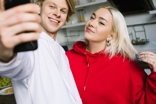 Friends taking selfie with cell phone. Posing. Red and white clothes hoodie. Casual style. At the kitchen in studio apartment