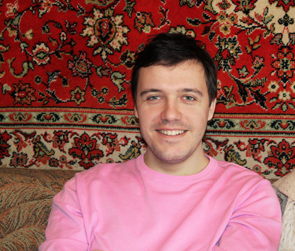 young man in pink jacket sits in traditional Soviet interior in style of 80s with carpet on wall visiting his grandmother