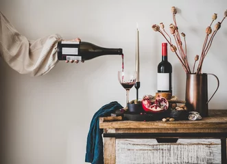 Foto op Plexiglas Red wine, board with cheese, fruit, almonds and olives, candles, decorative flowers on kitchen counter and womans hand pouring wine to glass, white wall at background. Winery, wine tasting concept © sonyakamoz