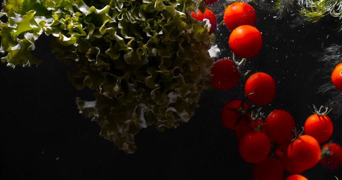 
A group of many ripe cherry tomatoes, bunches of dill and lettuce fall into the water on a black background with dramatic light, creating much bubbles. Slow motion 50 FPS 4K