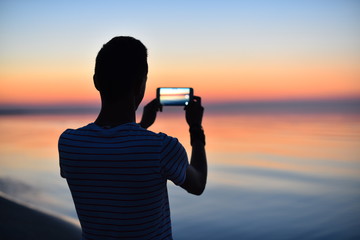 Silhouette of a guy taking photo with his mobile phone camera of a beautiful sunrise over the calm sea.. - obrazy, fototapety, plakaty