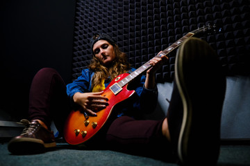 Obraz premium Female guitarist sitting on the floor in a recording Studio. A woman plays music on a red electric guitar. Denim jacket, cap, jeans and sneakers.
