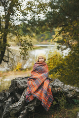 Little girl wrapped in a blanket is sitting on wooden stub near lake in an autumn forest