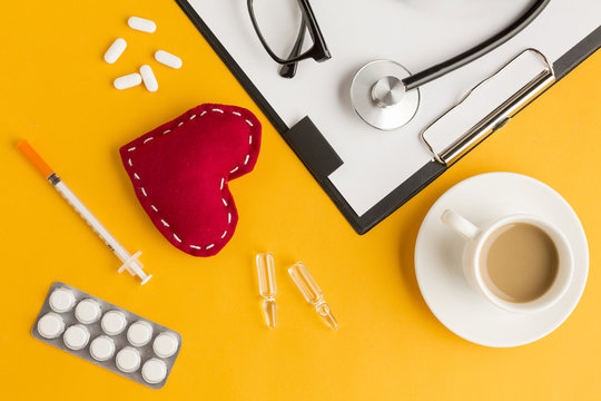 Red stitched heart; coffee cup; stethoscope; eyeglass; injection; blister packed medicine; clipboard against yellow backdrop