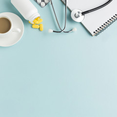 Tablets falling from bottle with coffee cup; blister packed tablets; stethoscope; spiral notepad against blue background
