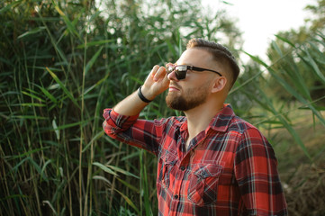 Outdoors portrait of young bearded man with sunglasses looking at the camera in the park on trees background