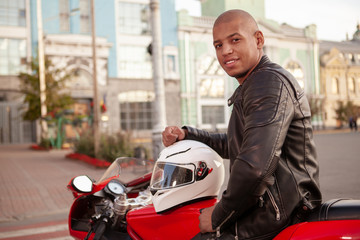 Fototapeta na wymiar Handsome African man in protective motorcycle jacket and jeans smiling to the camera, sitting on a motorbike. Cheerful male biker resting after riding motorcycle in the city