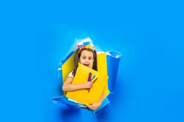 Child girl with book and bag breaking through blue paper wall.