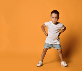 Kid boy in white t-shirt and jeans shorts is standing leaning forward with his hands on his hips at...