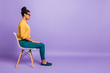 Fototapeta na wymiar Full length profile photo of pretty dark skin lady on chair look empty space listen employer question interview wear specs yellow shirt trousers isolated purple color background