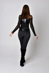 full length portrait of a pretty brunette woman wearing black leather fantasy costume with a dagger. standing pose, facing away from camera on a studio background.