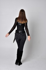 full length portrait of a pretty brunette woman wearing black leather fantasy costume with a dagger. standing pose, facing away from camera on a studio background.