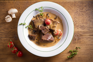 Braised beef cheeks in cream sauce with mushrooms and tomatoes in white bowl on wooden table. Overhead