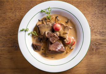 Braised beef cheeks in cream sauce with mushrooms and tomatoes in white bowl on wooden table. Overhead