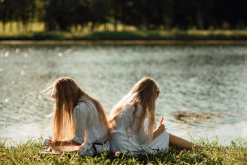 two little girls with pencil draws in notebook while sitting on the grass in the park, child writes in notebook