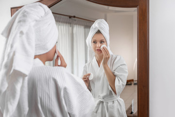 Young cute girl in a white waffle bathrobe with a towel on her head poses in a spacious white room looking in the mirror and rubbing her face with a cotton pad. Beauty and facial concept.