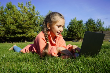 A girl of seven years old works on a laptop on the green grass. Distance learning. Girl with a laptop on a background of pines. Girl with a computer on the street.