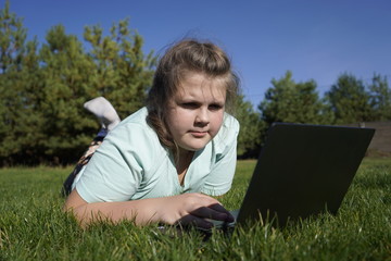 Teenage girl  working on a laptop on green grass. Distance learning. Girl with a laptop on a background of pines. Girl with a computer on the street.