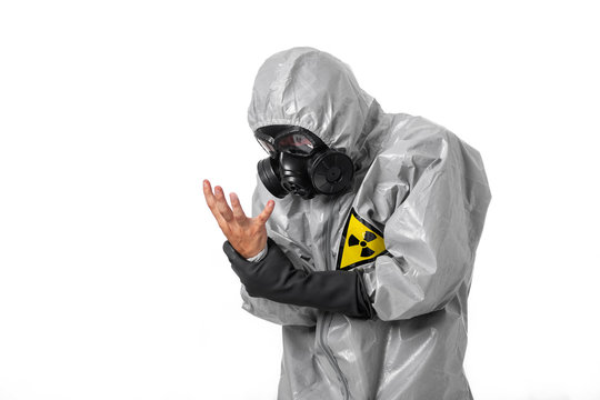 A man poses in a gray protective suit with a hood on his head with a yellow radiation sign, with a protective gas mask, posing while standing on a white background, with his hand without a glove. 