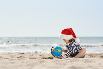 Obraz premium Happy little child girl in Santa hat sits on sand with globe on tropical beach. Kid having fun on summer vacation on Christmas and New year. Holiday Travel and adventure concept