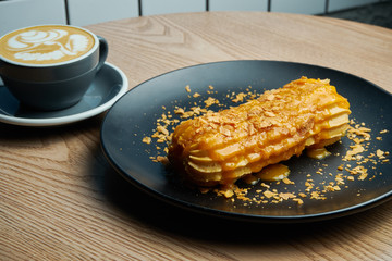 Appetizing and large eclair with glaze of mango on top and fir cream inside on a black plate. Sweets and dessert for coffee and tea.