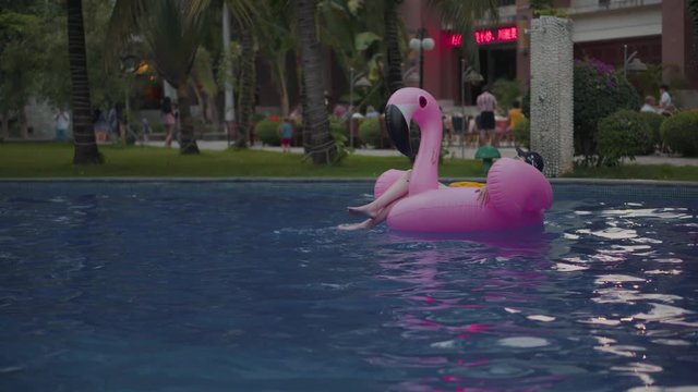 A young girl swims in the afternoon in the pool. Fashionable woman in a cap is rowing on a pink inflatable flamingo. Mattress, flamingo, girl in an open air hotel.