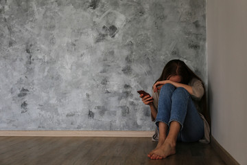 Portrait of beautiful young woman with depressed facial expression sitting in the corner holding her phone. Cyber bullying victim concept. Sad female on the floor of her room. Background, copy space. - obrazy, fototapety, plakaty