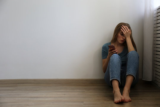 Portrait of beautiful young woman with depressed facial expression sitting in the corner holding her phone. Cyber bullying victim concept. Sad female on the floor of her room. Background, copy space.