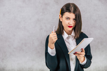 Cute business women standing with pencil behind her ear and listening, writing down something into the notebook, pointing pen at the camera.