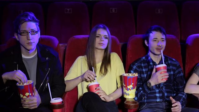 Young fashionable teenagers sit in the cinema and watch a Comedy or a melodrama, eat popcorn and drink Cola. Portrait of smiling teenagers in a modern cinema.