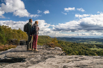 two hikers (couple - man and woman) up on the rock - looking into valley below mountain on forest...