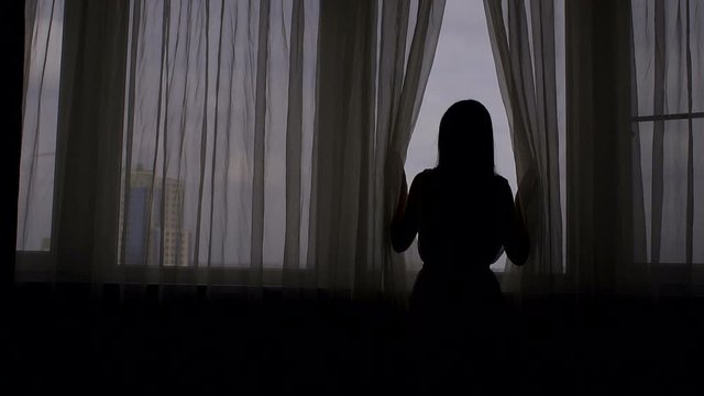 Silhouette. Beautiful woman opening curtains and looking through the window. Attractive sexy woman with slender body is looking at the sunrise standing near the window.