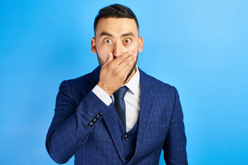 Frightened shocked Asian Kazakh man in blue suit and tie with bulging surprised eyes closes mouth...