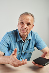 Authentic photo portrait of elderly talking man sitting at table with mobile phone in hand, modern advanced candid senior citizen on white background