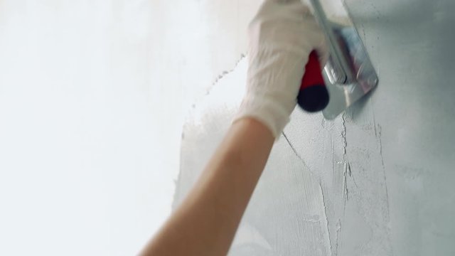 Close-up of female hands applying gray paint or plaster with a spatula on the wall.