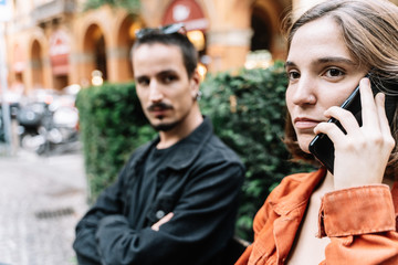 Girl talking to the mobile while a boy waits and looks at her with serious expression.