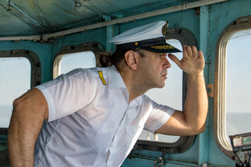 Captain standing in the wheelhouse and looking to ahead. A sailor in the white uniform looking to sea from control room - navigation bridge. - obrazy, fototapety, plakaty