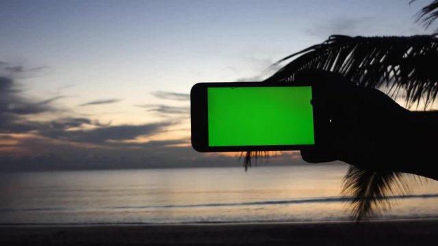 The man raised his hand on a green screen electronic device to record the seashore at sunset.