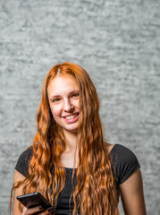 portrait of young teenager redhead girl with long hair with smartphone on gray wall background