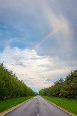 Small rainbow over trees on either side of a straight road